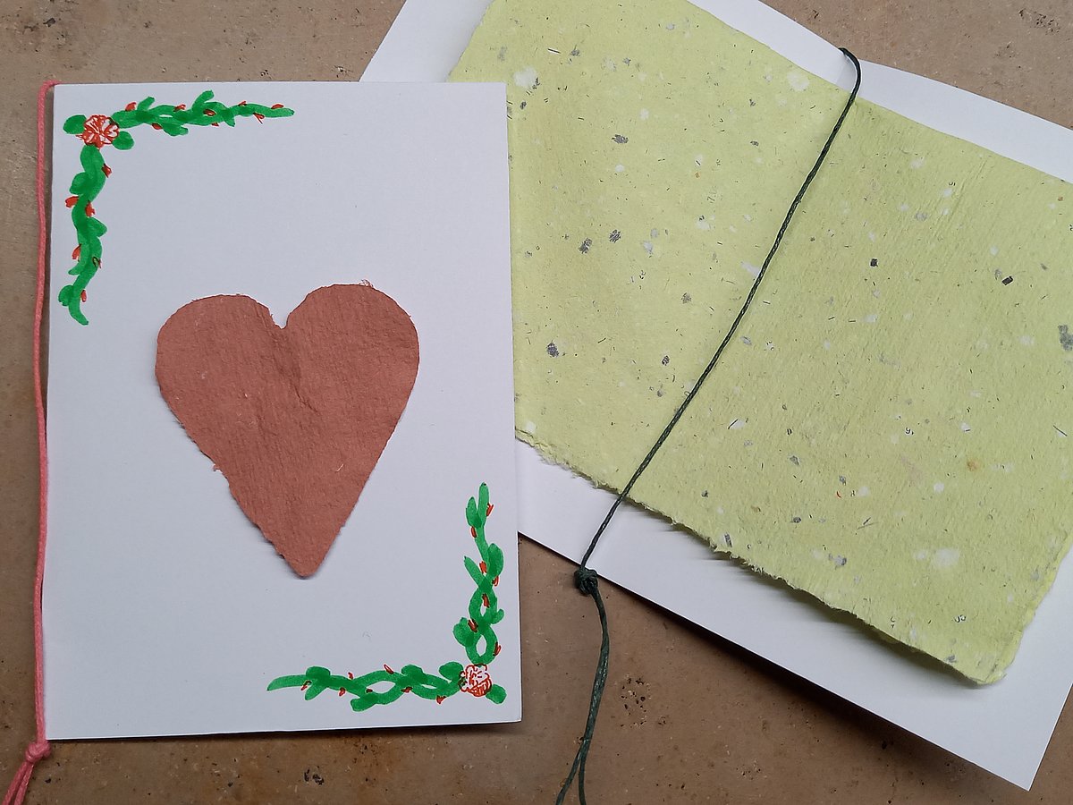 Zwei Karten aud weißem Karton wurden mit selbst geschöpftem, rauem Papier verziert. Eine Karte zeigt außen ein rotes Herz aus Papier und gezeichnete, grüne Ranken. In die zweite Karte wurde innen ein hellgrünes, geflecktes Stück selbst geschöpftes Papier gelegt und mit einem Faden festgebunden.